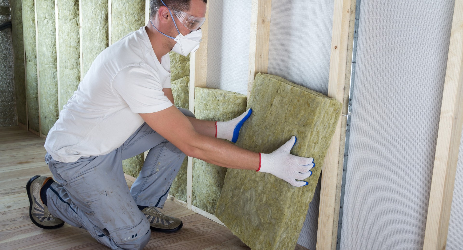 Worker in protective goggles and respirator insulating rock wool insulation in wooden frame for future house walls for cold barrier. Comfortable warm home, economy, construction and renovation concept