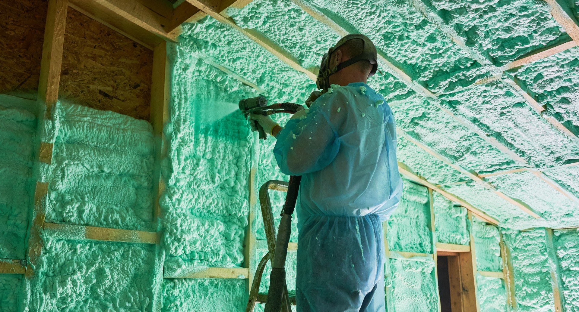 Male builder insulating wooden frame house. Man worker spraying polyurethane foam inside of future cottage, standing on ladder, using plural component gun. Construction and insulation concept.